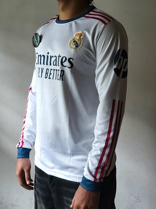A white long-sleeve Real Madrid soccer jersey with red stripes on the shoulders and sleeves, blue trim, and dark sponsor logos, shown on a model against a gray wall.