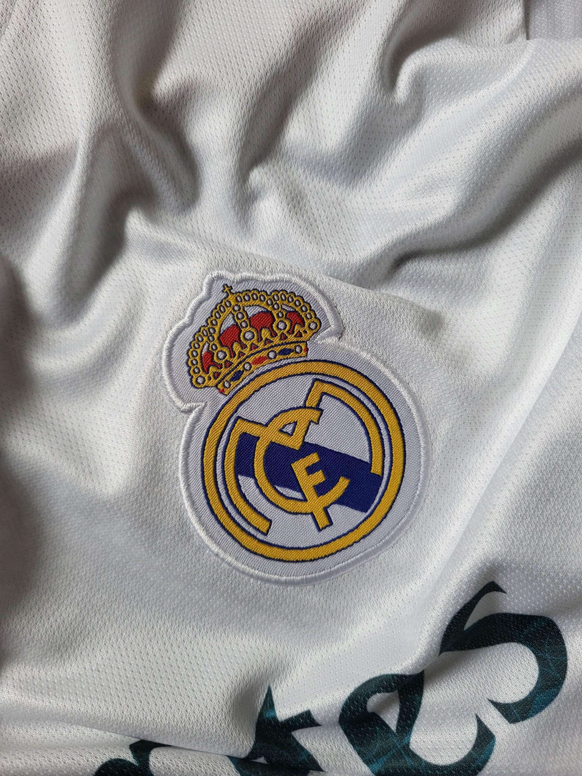 Close-up of a white Real Madrid soccer jersey fabric showing the embroidered club crest and a small gold-and-red crown patch on the chest.