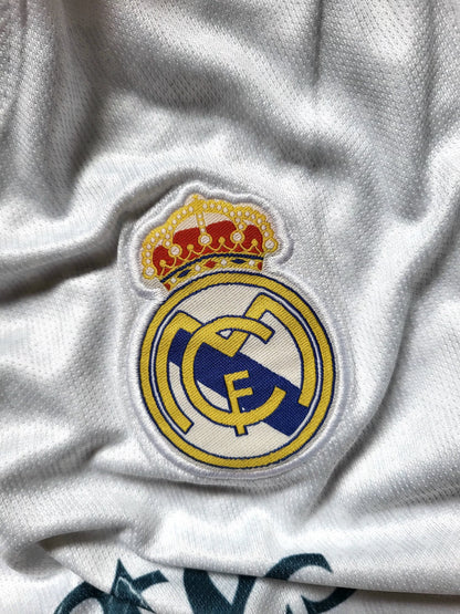 Close-up of a white Real Madrid soccer jersey showing the embroidered club crest with a gold crown on the chest.