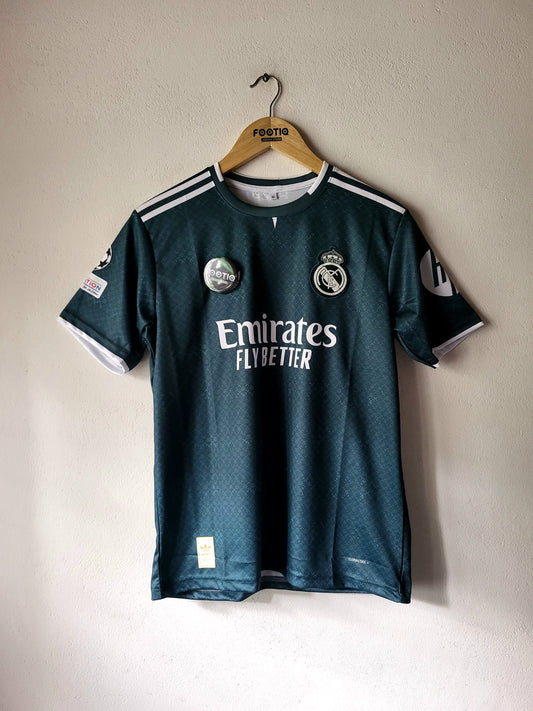 A dark teal Real Madrid soccer jersey hanging on a wooden hanger against a plain beige wall. The jersey features white 'Emirates Fly Better' sponsor text across the chest, the Real Madrid club crest, and white shoulder stripes.