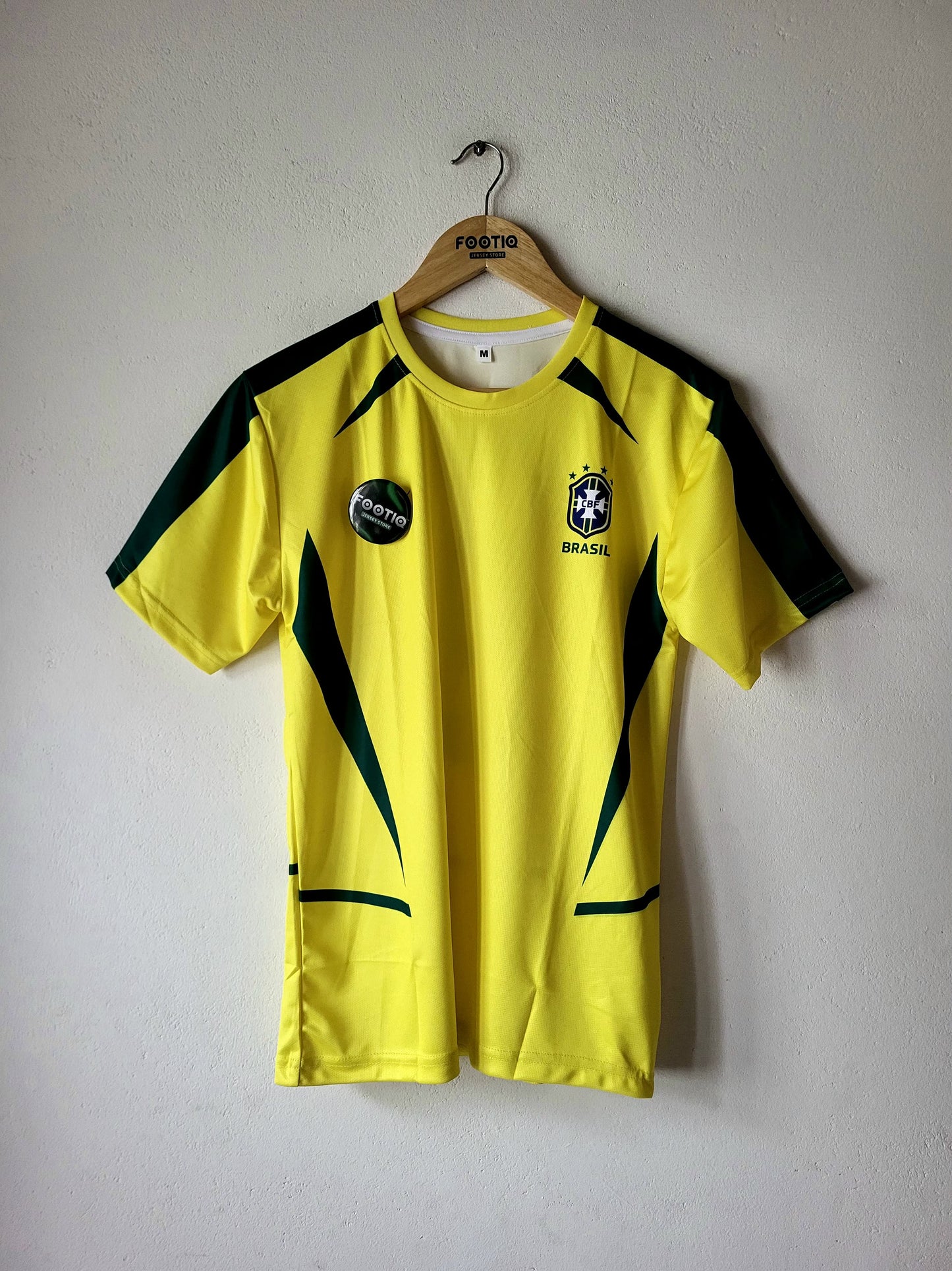 A yellow Brazil soccer jersey with green accents hanging on a wooden hanger against a light gray wall. The jersey features the Brazilian national team crest on the left chest and a circular patch on the right chest.