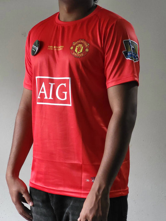 A red Manchester United soccer jersey with the AIG sponsor logo on the front, worn by a model against a plain gray background.