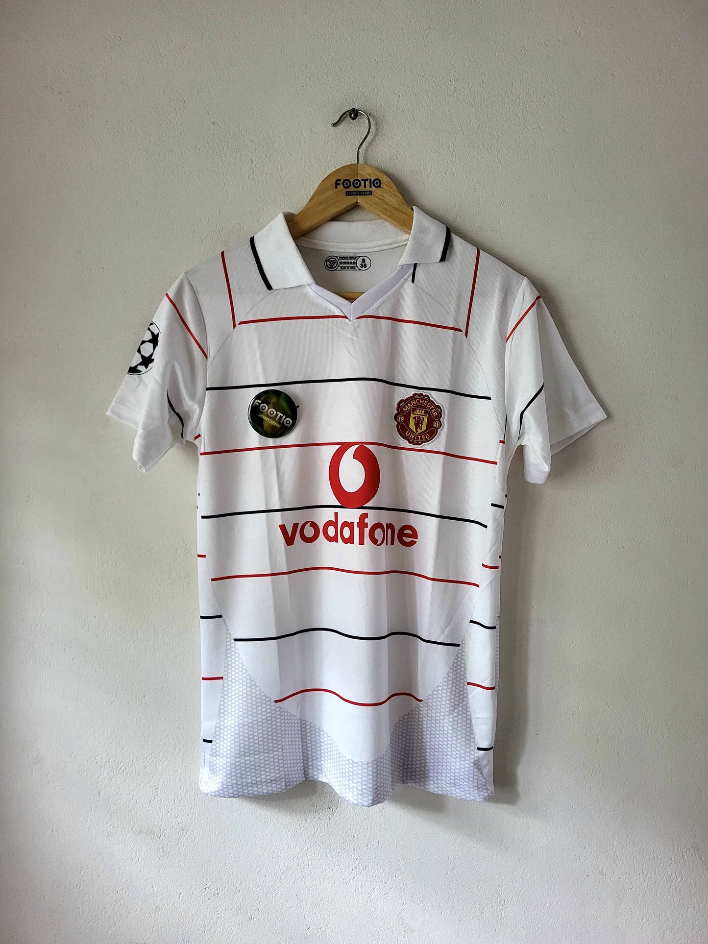 A vintage white Manchester United football shirt with red and black stripes, featuring the red Vodafone logo and club crest, hanging on a wooden hanger against a plain wall.