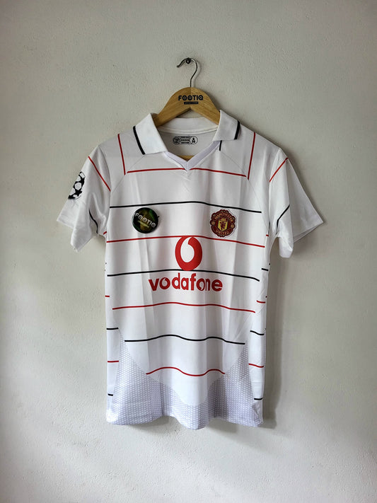 A vintage white Manchester United football shirt with red and black stripes, featuring the red Vodafone logo and club crest, hanging on a wooden hanger against a plain wall.