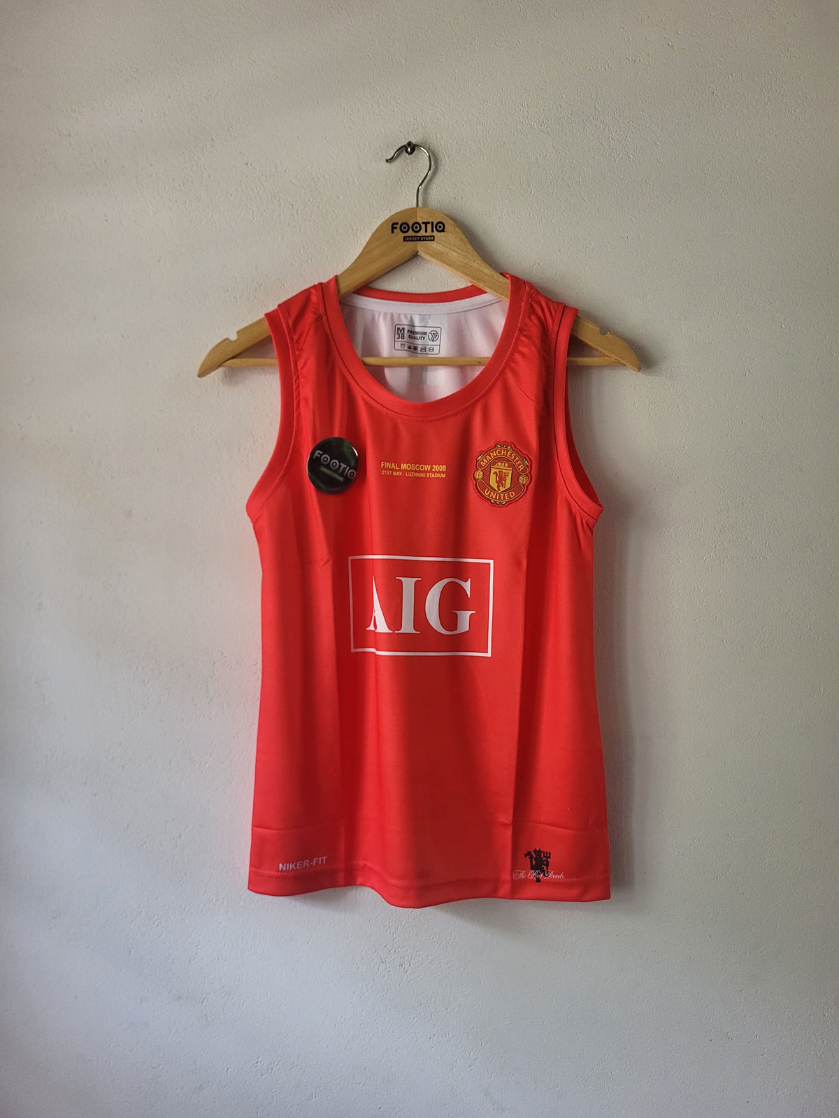 A red sleeveless Manchester United athletic jersey hanging on a wooden hanger against a plain light gray wall. The jersey features the Manchester United crest on the left chest, a large white 'AIG' sponsor logo on the front, and a small black circular patch on the right chest.
