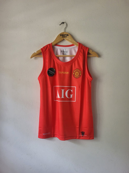 A red sleeveless Manchester United athletic jersey hanging on a wooden hanger against a plain light gray wall. The jersey features the Manchester United crest on the left chest, a large white 'AIG' sponsor logo on the front, and a small black circular patch on the right chest.