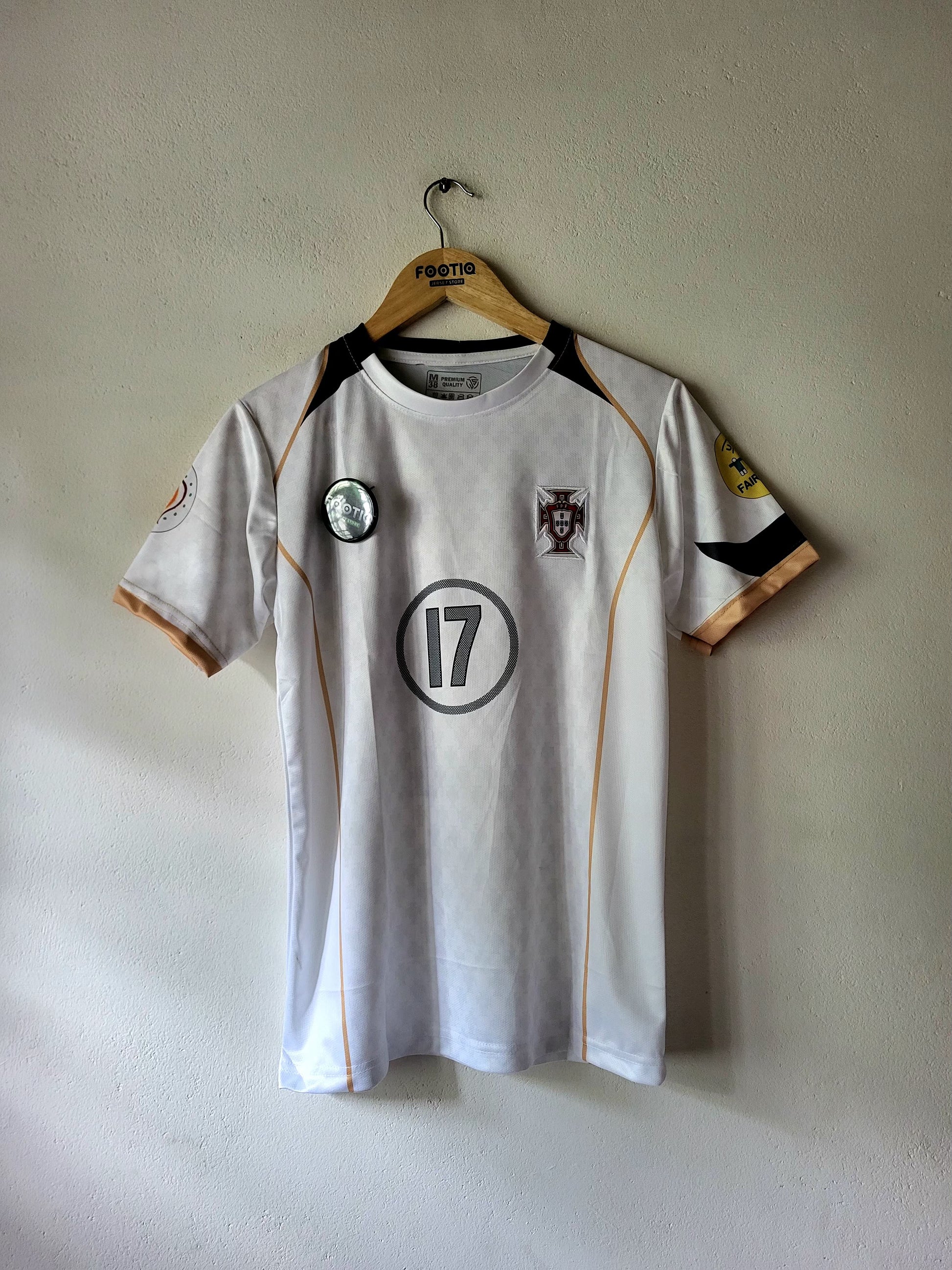 A white Portugal soccer jersey with black and gold accents, featuring the number 17 on the chest, hanging on a wooden hanger against a textured beige wall.