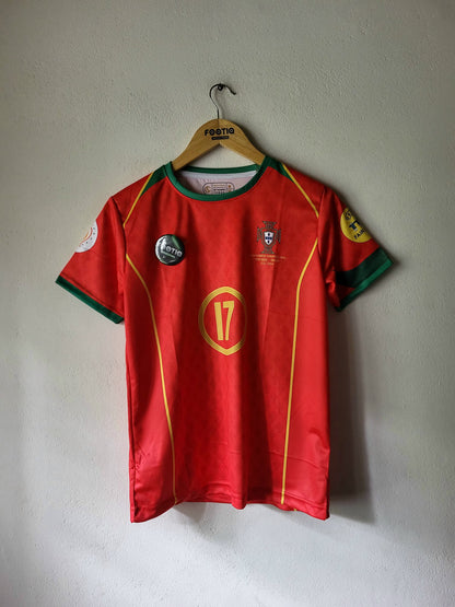 A red Portugal national football team jersey with green and yellow trim, hanging on a wooden hanger against a textured white wall. The jersey features the number 17 on the front, the Portugal crest, and various sponsor patches.