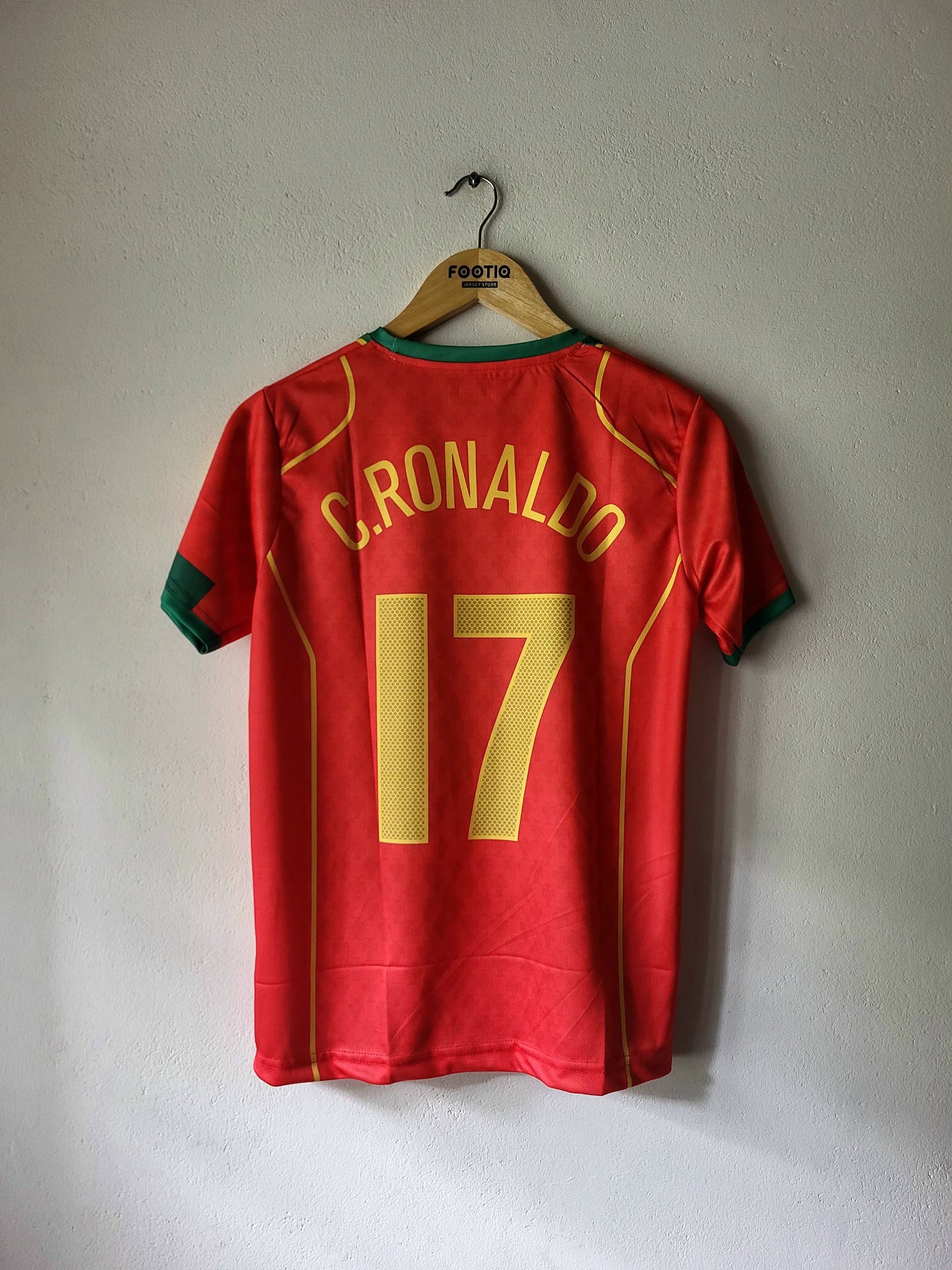 A red Portugal soccer jersey with 'C. RONALDO' and the number '17' in yellow on the back, hanging on a wooden hanger against a light gray wall.