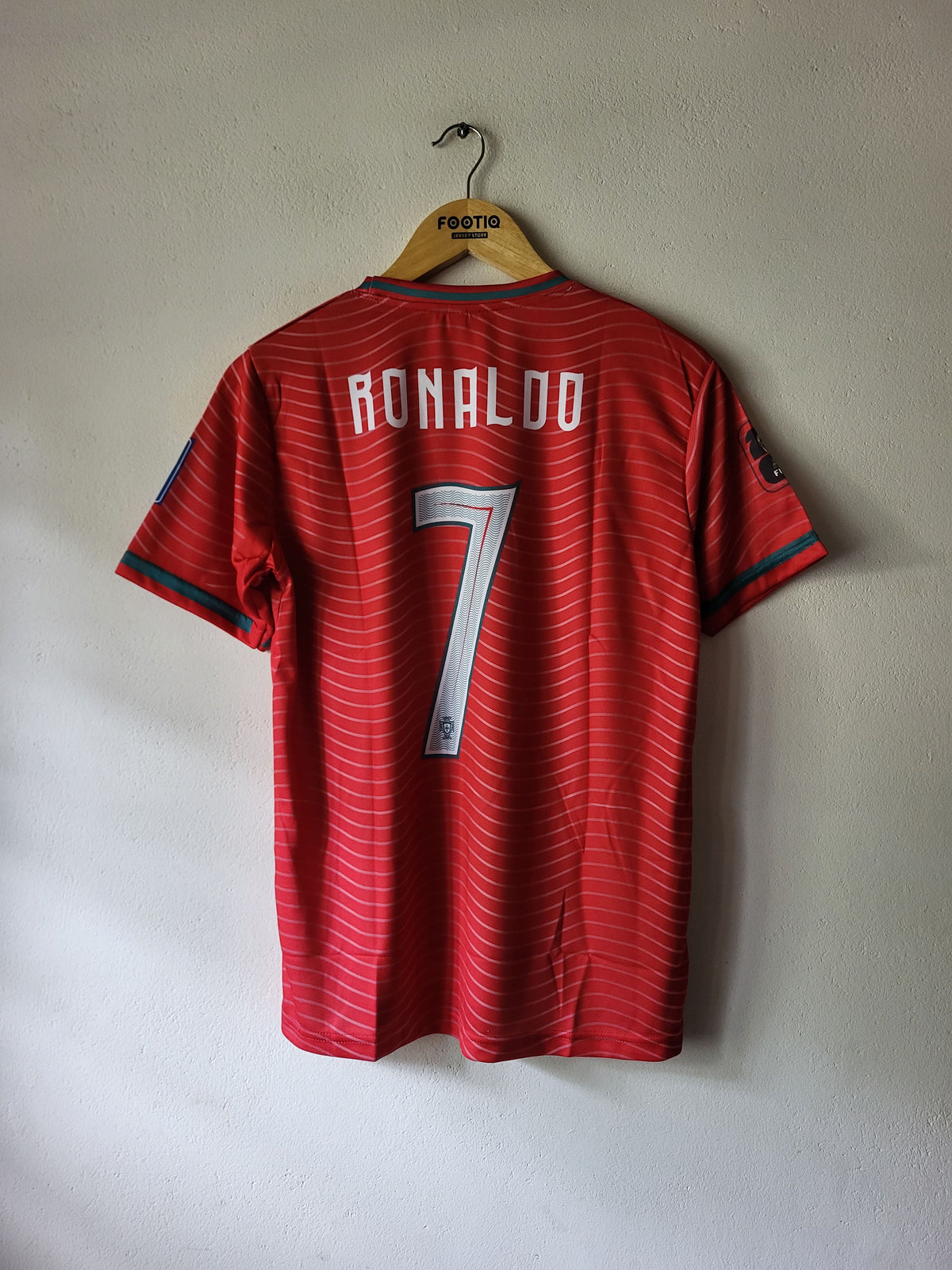 A red short-sleeve soccer jersey with 'RONALDO 7' printed on the back, hanging on a wooden hanger against a plain light gray wall.