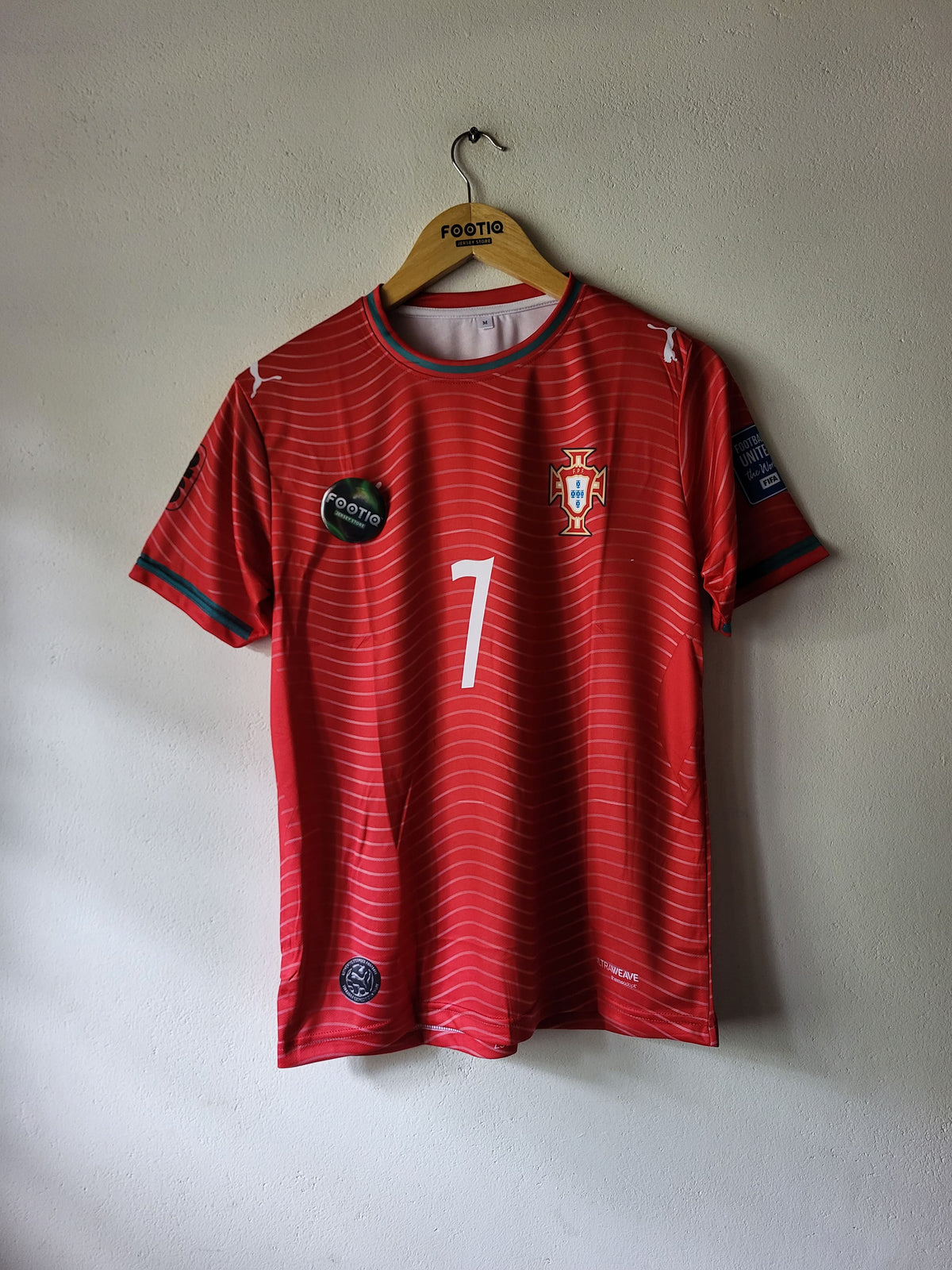 A red Portugal soccer jersey with the number 7 on the front, hanging on a wooden hanger against a textured off-white wall.