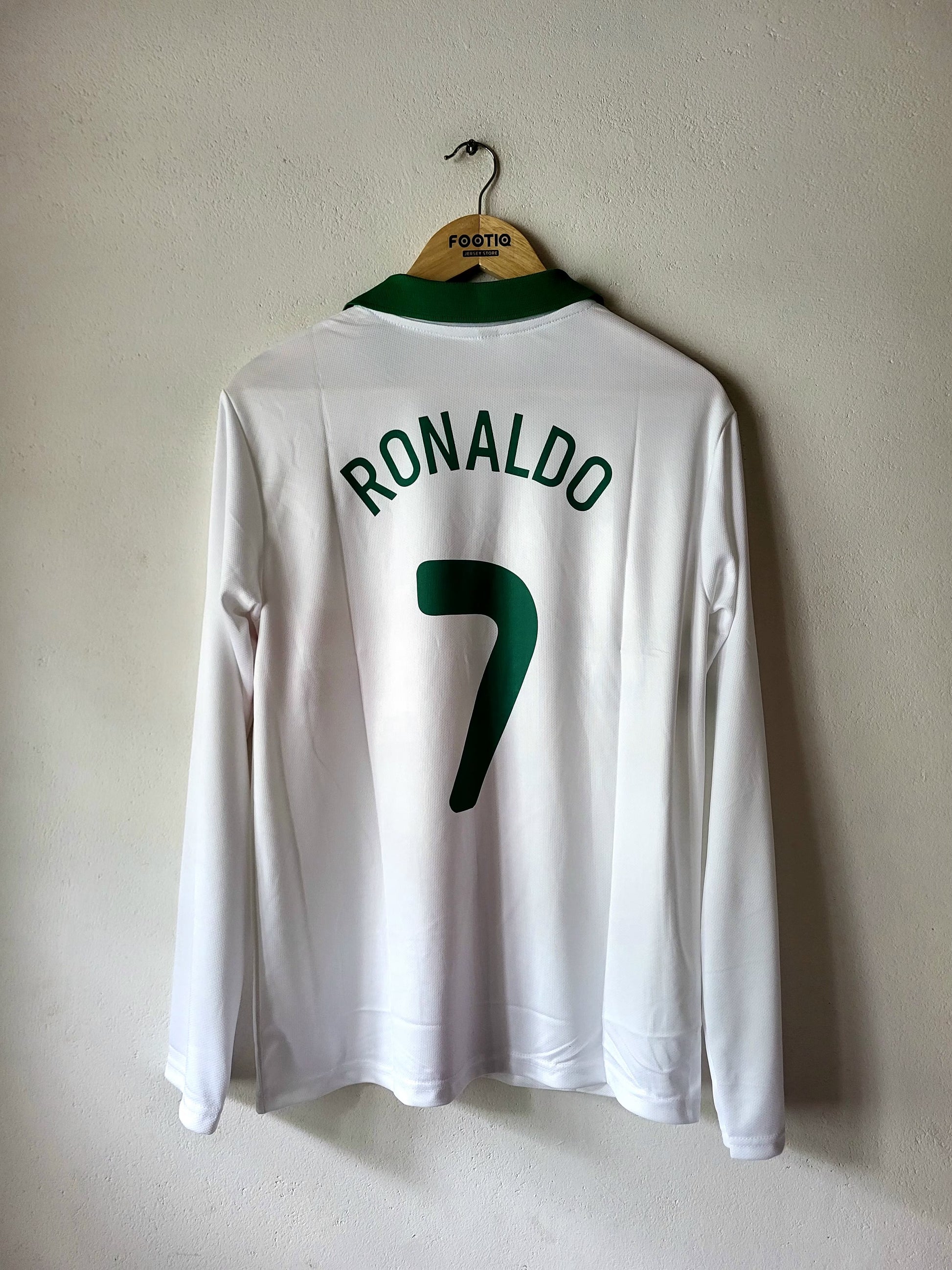 A white long-sleeve soccer jersey hanging on a wooden hanger against a plain beige wall. The back of the jersey has 'RONALDO' and the number '7' printed in dark green, with a green collar.