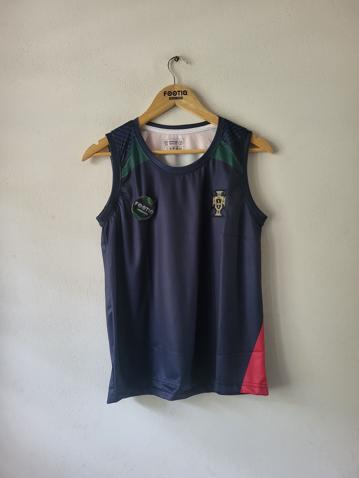 A sleeveless athletic tank top hanging on a wooden hanger against a plain off-white wall. The top is dark navy blue with green panels on the upper shoulders, a gold Portugal crest on the left chest, a circular green patch on the right chest, and a red panel on the lower right side.