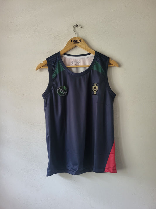 A sleeveless athletic tank top hanging on a wooden hanger against a plain off-white wall. The top is dark navy blue with green panels on the upper shoulders, a gold Portugal crest on the left chest, a circular green patch on the right chest, and a red panel on the lower right side.