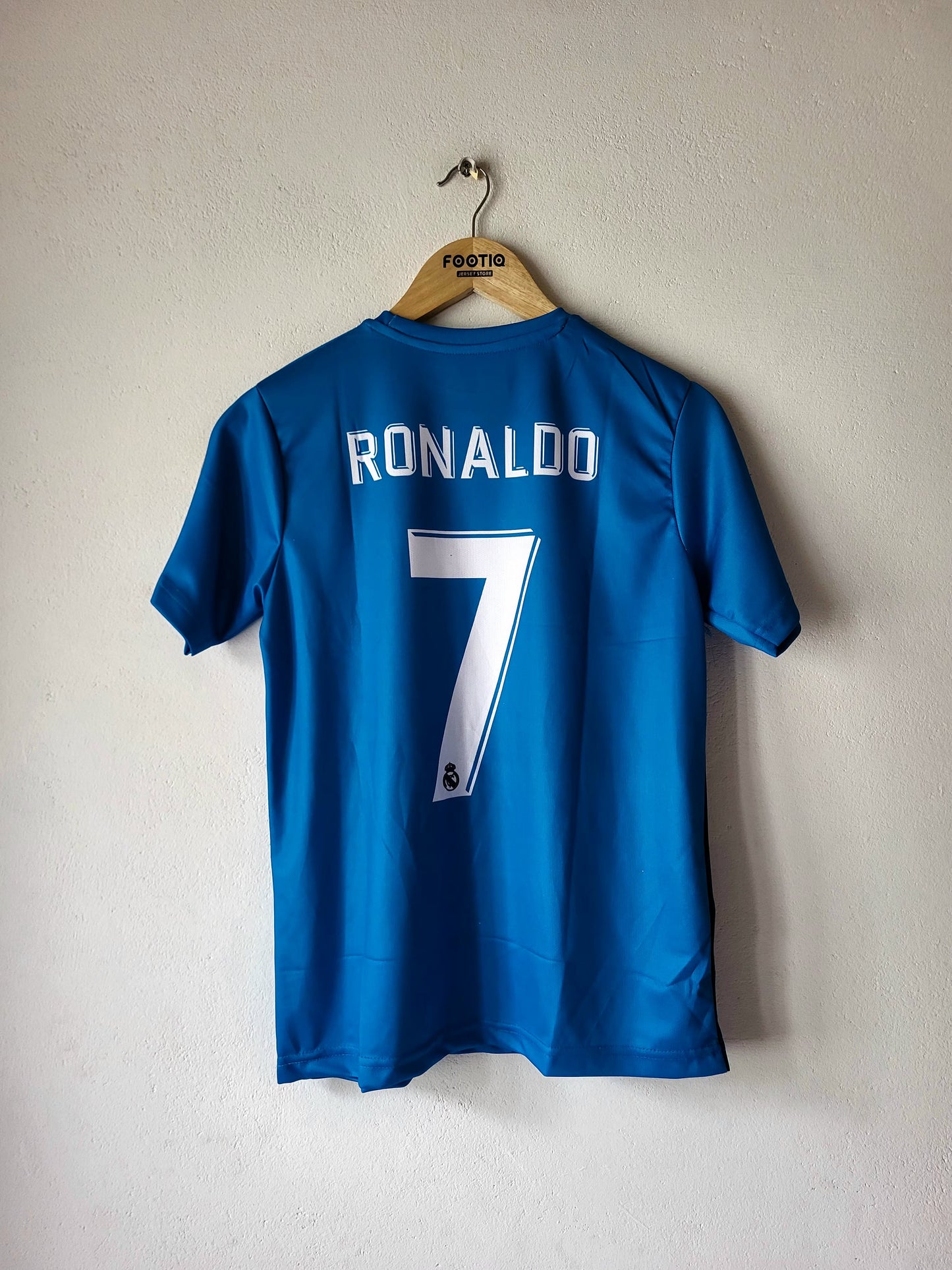A blue soccer jersey with 'RONALDO' and the number '7' printed in white on the back, hanging on a wooden hanger against a textured off-white wall.