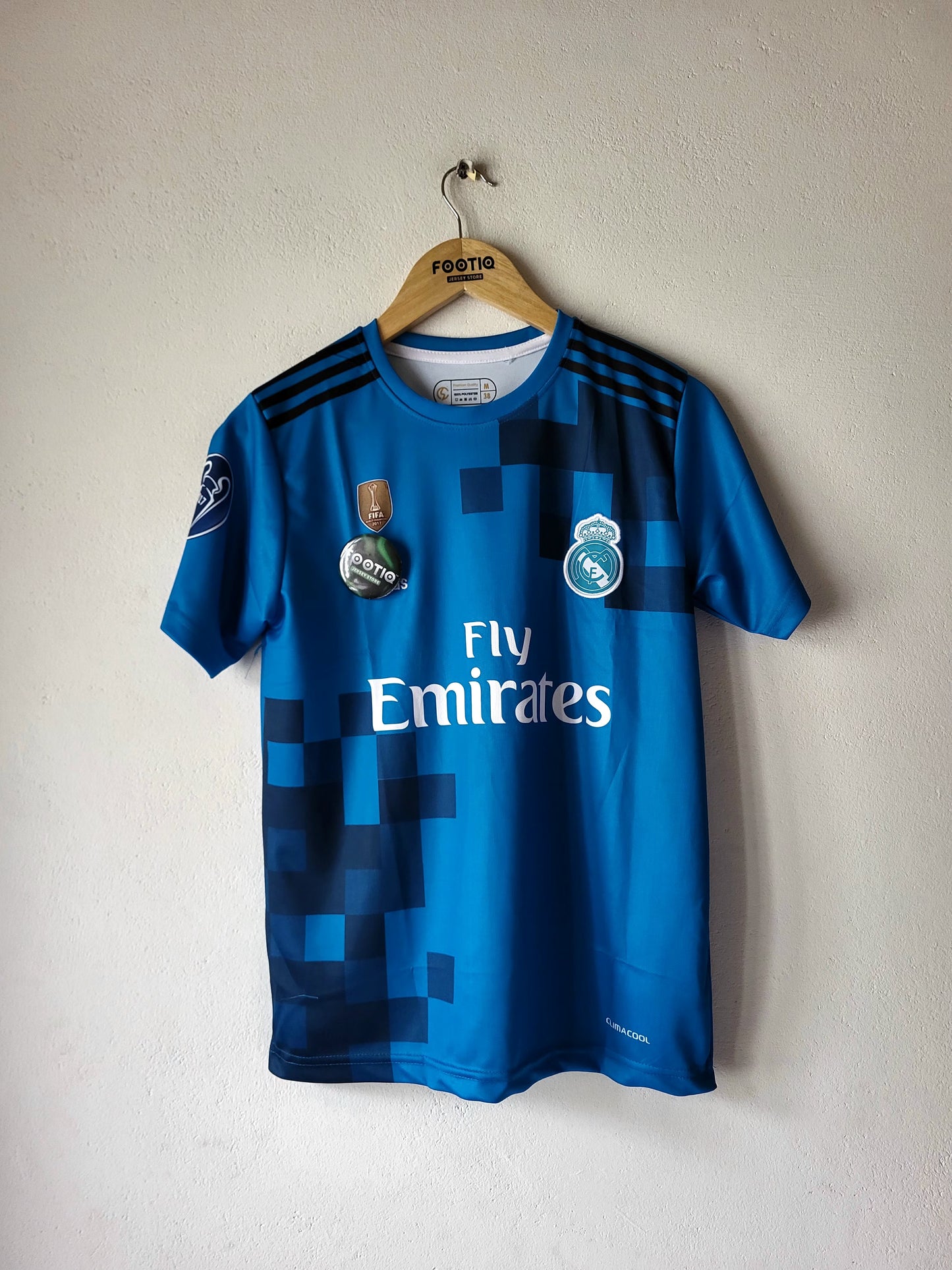 A blue Real Madrid soccer jersey with pixelated dark blue patterns, hanging on a wooden hanger against a textured off-white wall. The jersey features the 'Fly Emirates' sponsor logo on the front, the club crest on the right chest, and the CFP badge on the left.