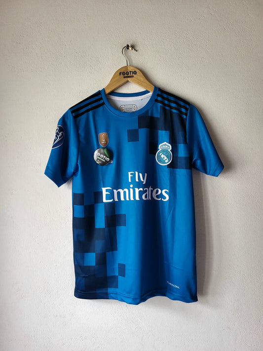 A blue Real Madrid soccer jersey with pixelated dark blue patterns, hanging on a wooden hanger against a textured off-white wall. The jersey features the 'Fly Emirates' sponsor logo on the front, the club crest on the right chest, and the CFP badge on the left.