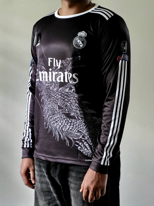 A person wearing a black long-sleeve Real Madrid soccer jersey with white stripes, a large white dragon graphic, and 'Fly Emirates' text, paired with dark jeans against a plain light background.