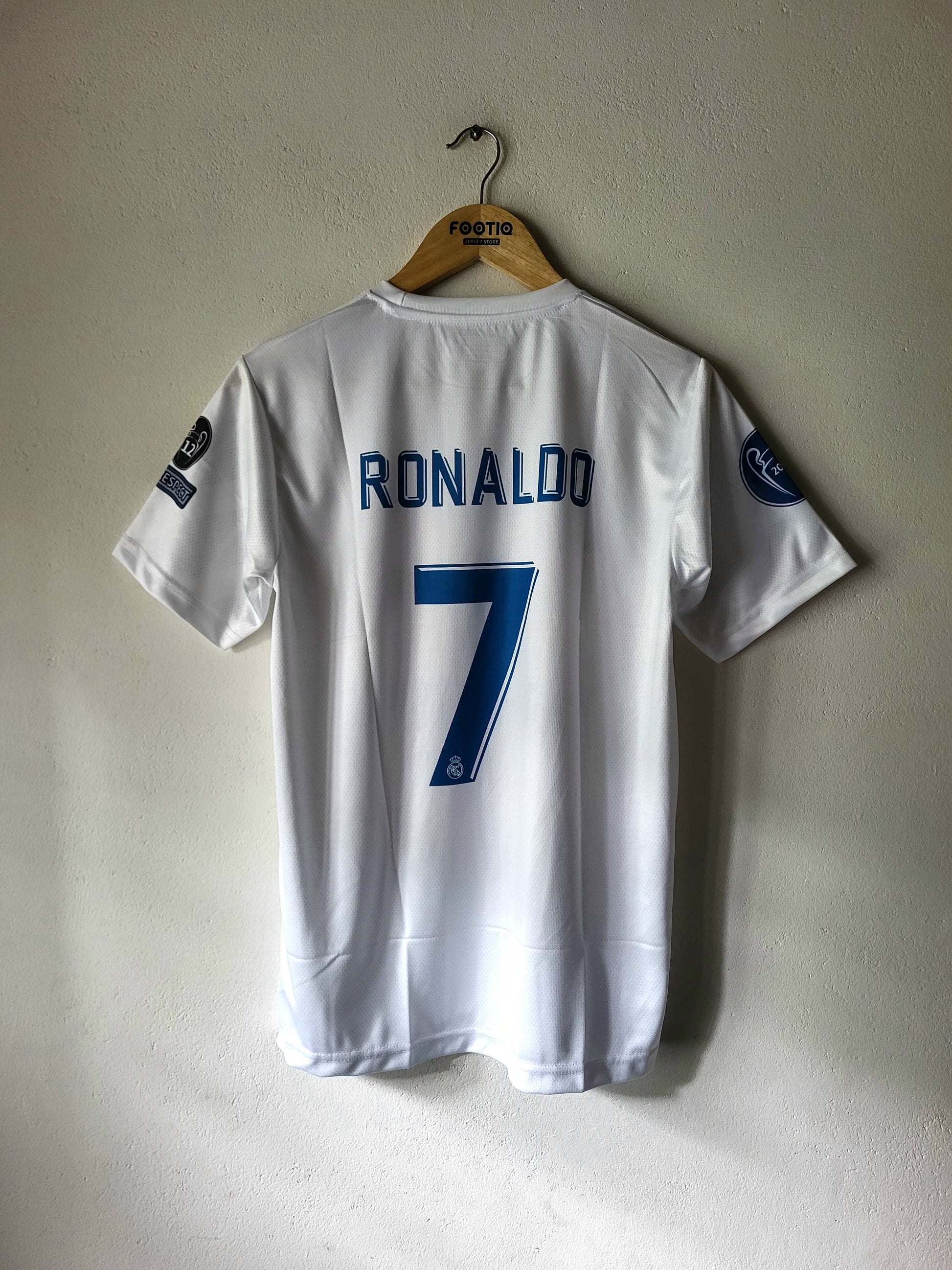 A white Sporting CP soccer jersey with 'RONALDO' and the number '7' printed in blue on the back, hanging on a wooden hanger against a textured off-white wall.