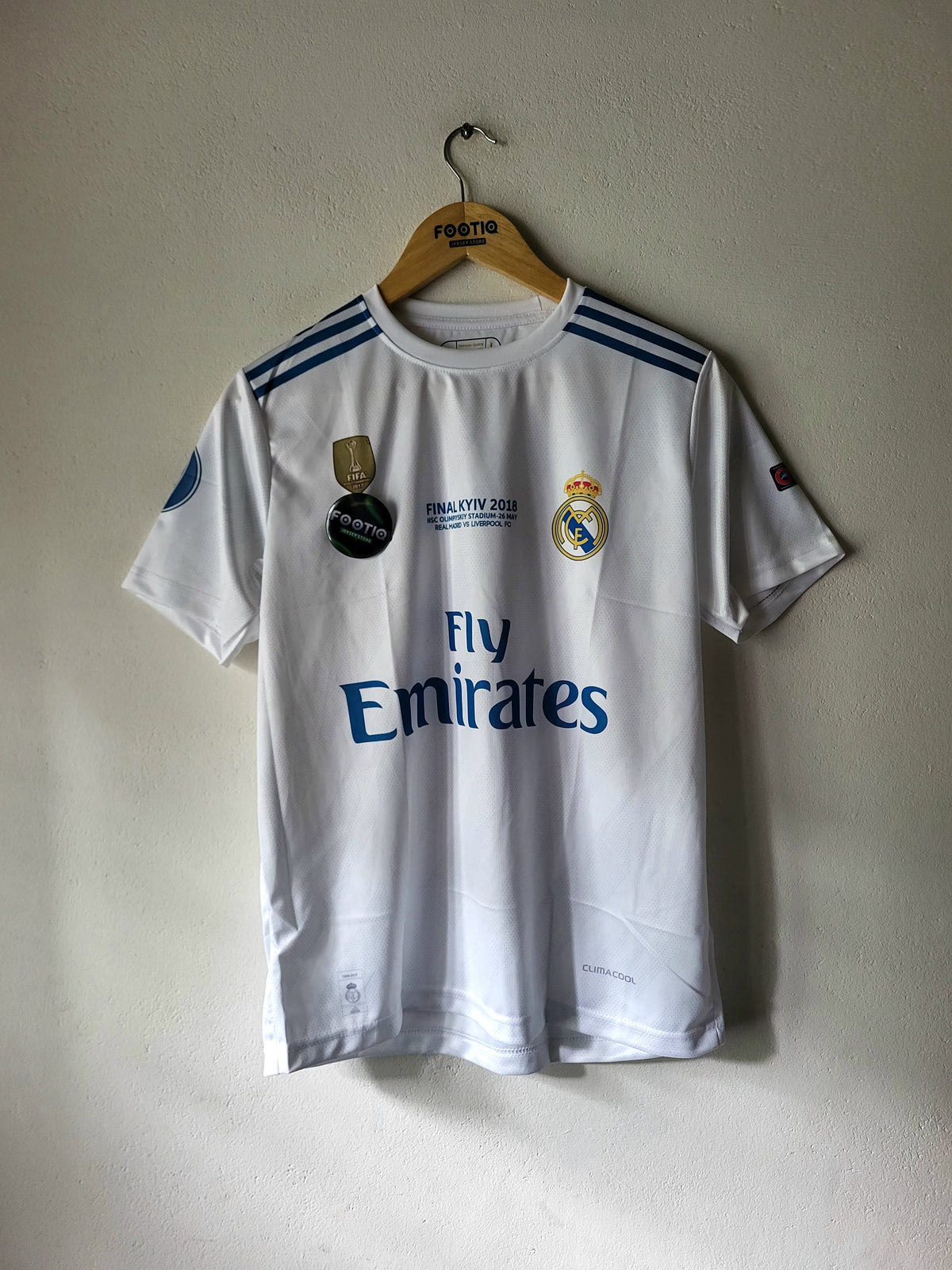 A white Real Madrid 2017/18 home soccer jersey hanging on a wooden hanger against a textured off-white wall. The jersey features blue shoulder stripes, the Real Madrid crest, the 'Fly Emirates' sponsor logo, and a 'Football 2017' patch on the chest.