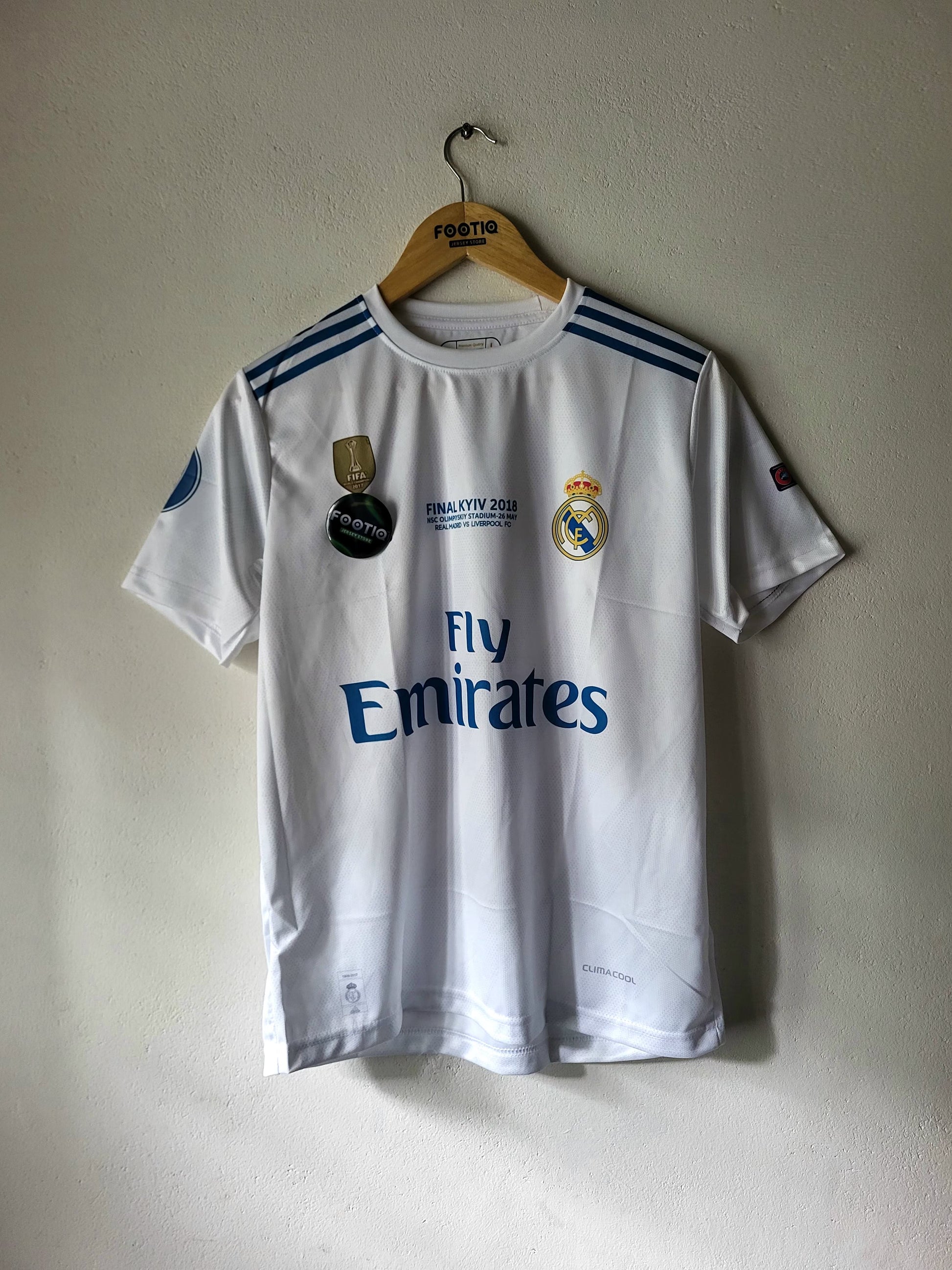 A white Real Madrid 2017/18 home soccer jersey hanging on a wooden hanger against a textured off-white wall. The jersey features blue shoulder stripes, the Real Madrid crest, the 'Fly Emirates' sponsor logo, and a 'Football 2017' patch on the chest.