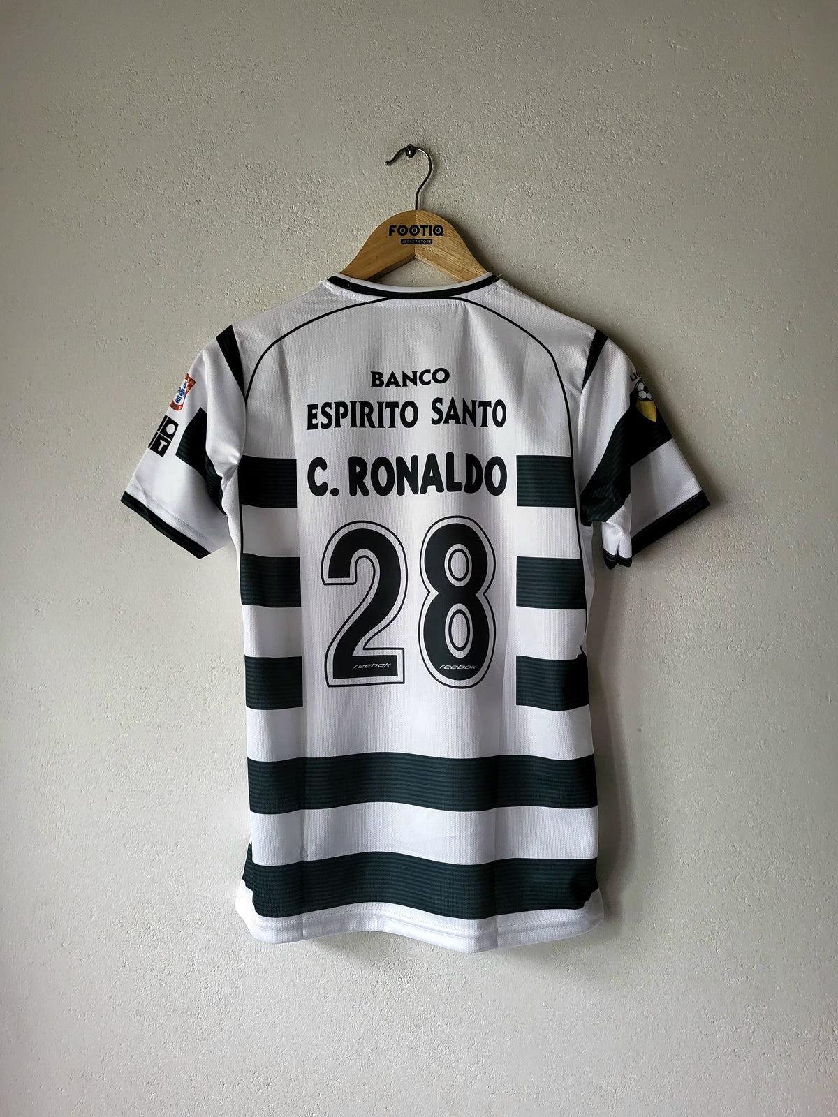 A white and dark green striped soccer jersey with 'C. RONALDO 28' printed on the back, hanging on a wooden hanger against a plain wall.