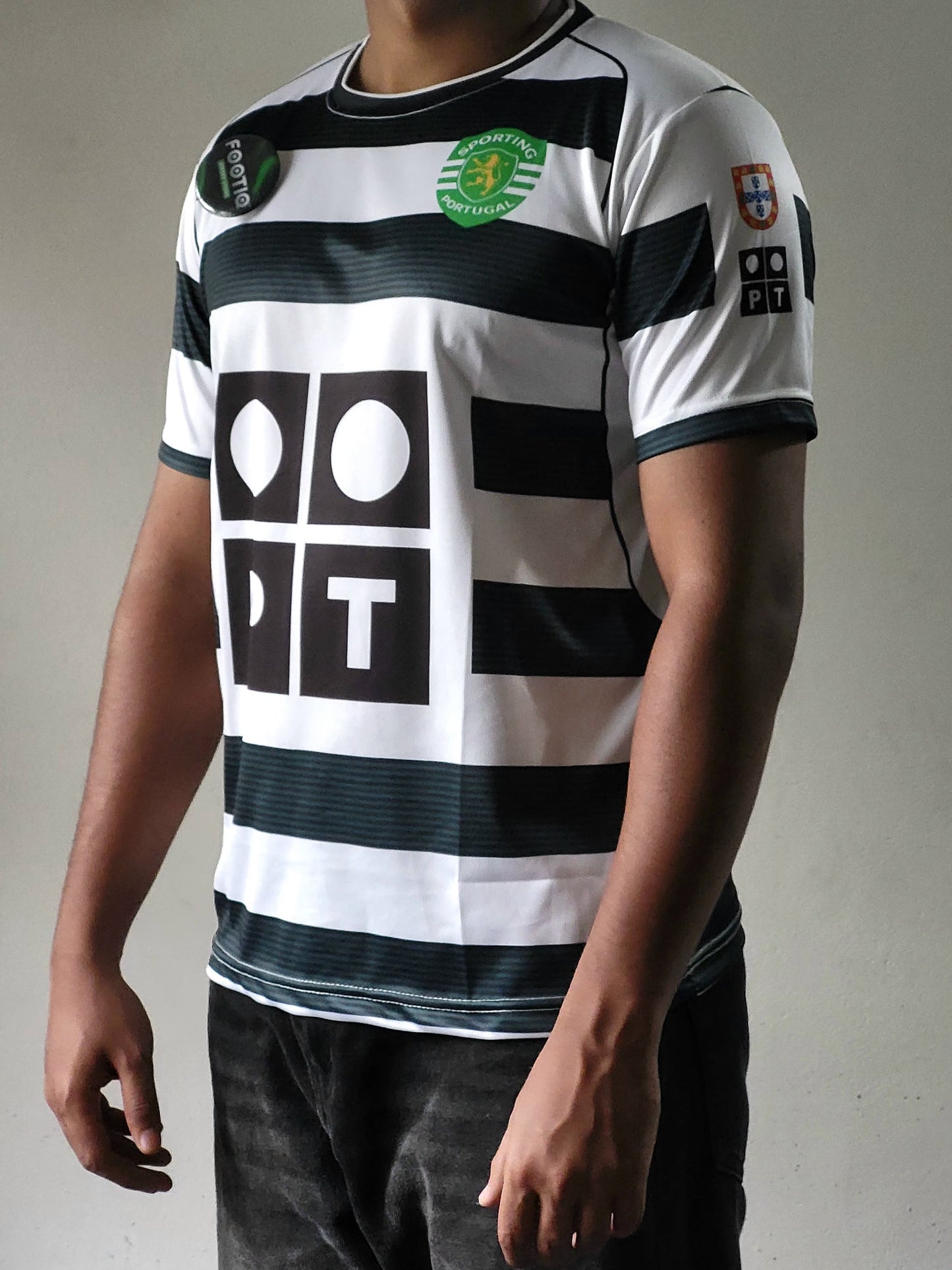 A man wearing a white and black horizontally striped soccer jersey with a green and yellow Sporting Portugal crest on the chest, a large black geometric logo on the front, and a Portugal national team patch on the sleeve.