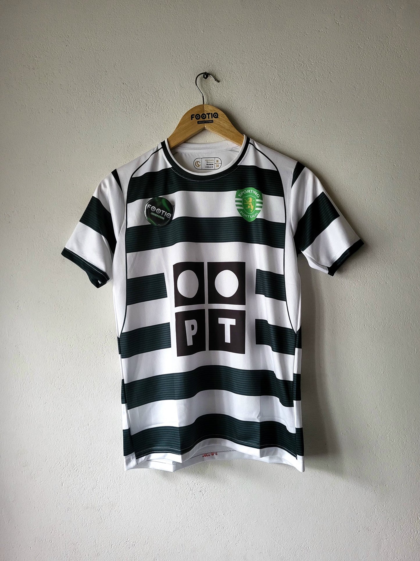 A black and white horizontally striped soccer jersey hanging on a wooden hanger against a textured gray wall. The jersey features a green and white crest on the left chest, a black circular patch on the right chest, and a large black 'PT' logo with white circles in the center.