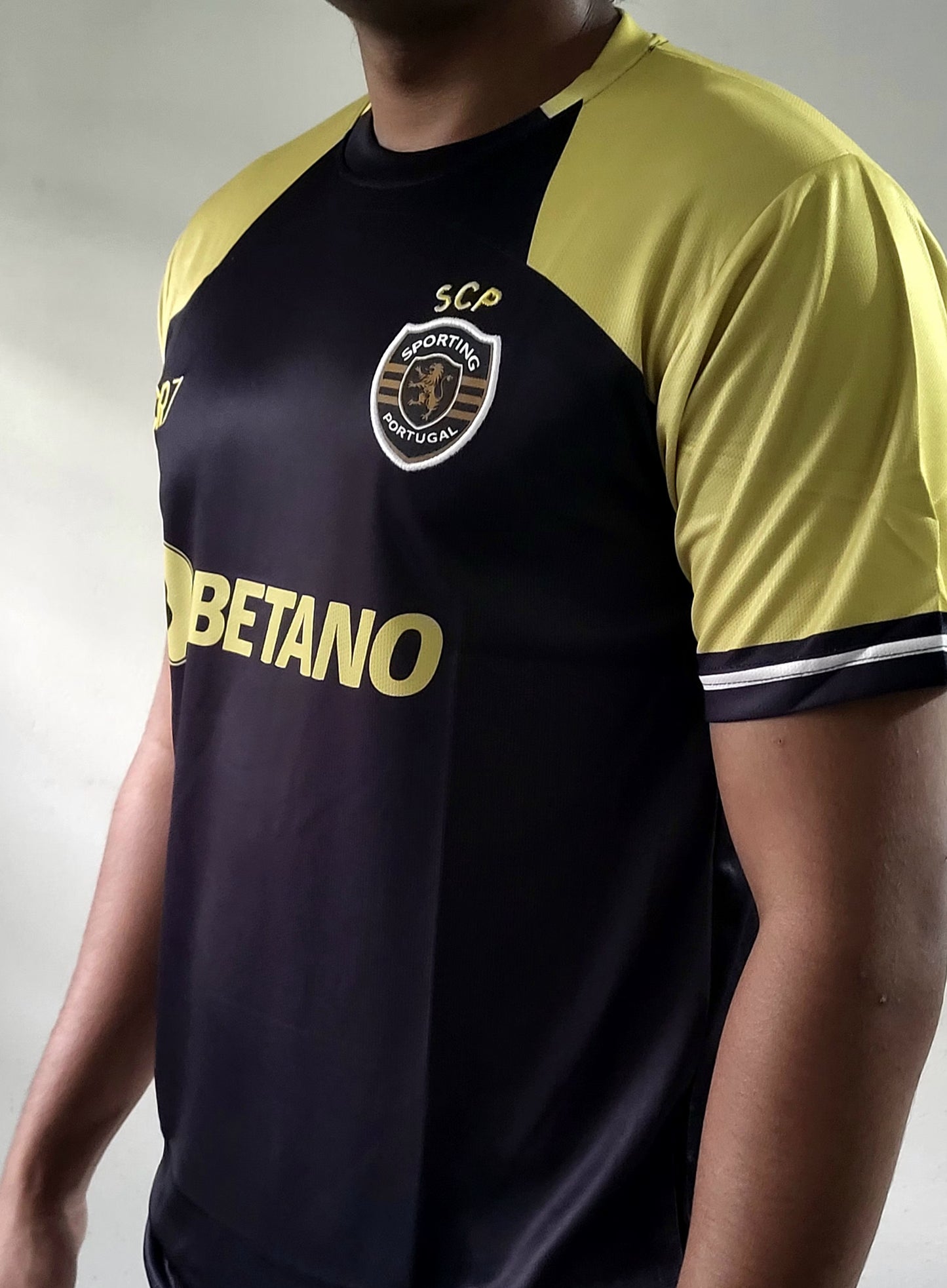A close-up of a soccer jersey worn by a model, featuring a black body with yellow shoulder panels, a yellow 'BETANO' sponsor logo, and the Sporting CP crest on the chest.