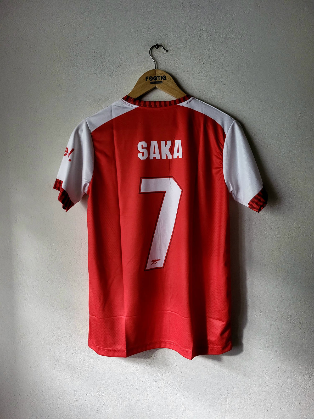 A red and white short-sleeve soccer jersey hanging on a wooden hanger against a light gray wall. The back of the jersey has 'SAKA' and the number '7' printed in white, with a small red AS United club crest near the bottom.