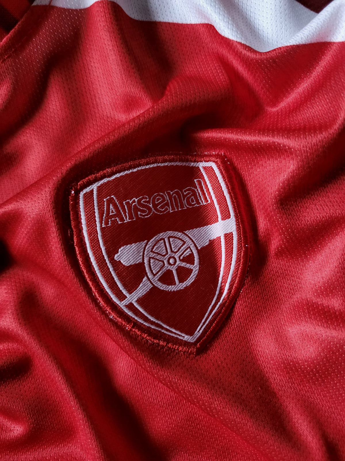 Close-up of a red Arsenal football club crest patch embroidered on a red and white satin-textured jersey fabric.