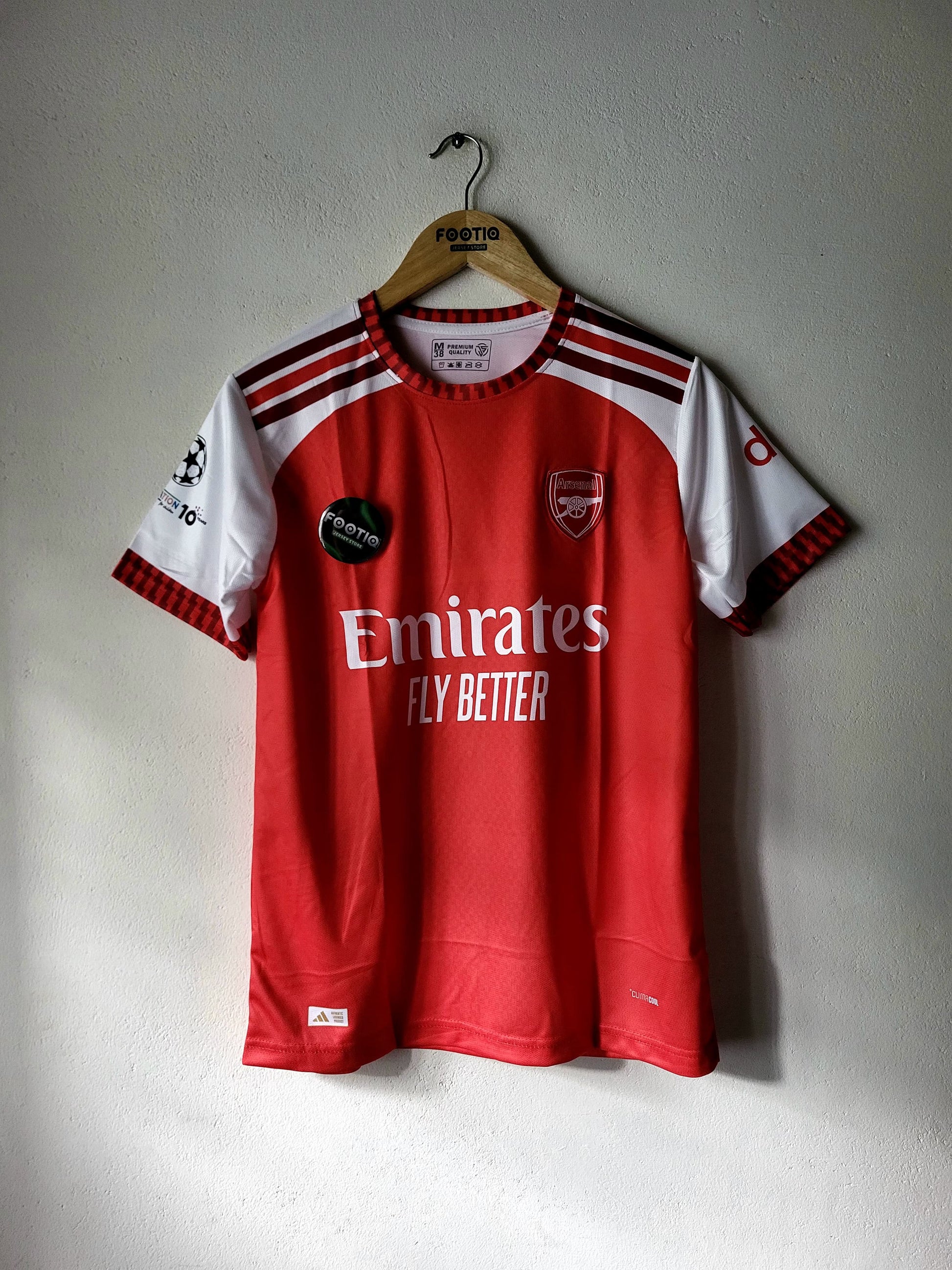 A red and white Arsenal soccer jersey hanging on a wooden hanger against a textured gray wall. The jersey features the Emirates Fly Better sponsor logo, the Arsenal club crest, and a UEFA Champions League patch on the sleeve.