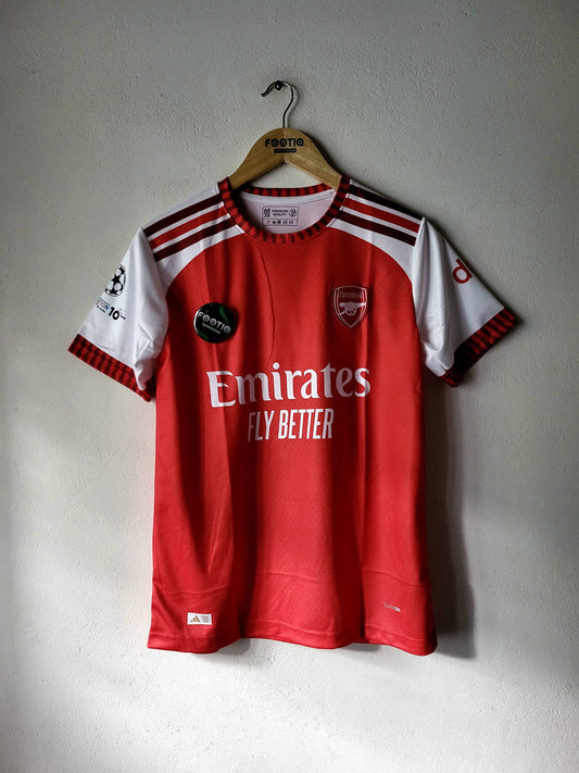 A red and white Arsenal soccer jersey hanging on a wooden hanger against a textured gray wall. The jersey features the Emirates Fly Better sponsor logo, the Arsenal club crest, and a UEFA Champions League patch on the sleeve.