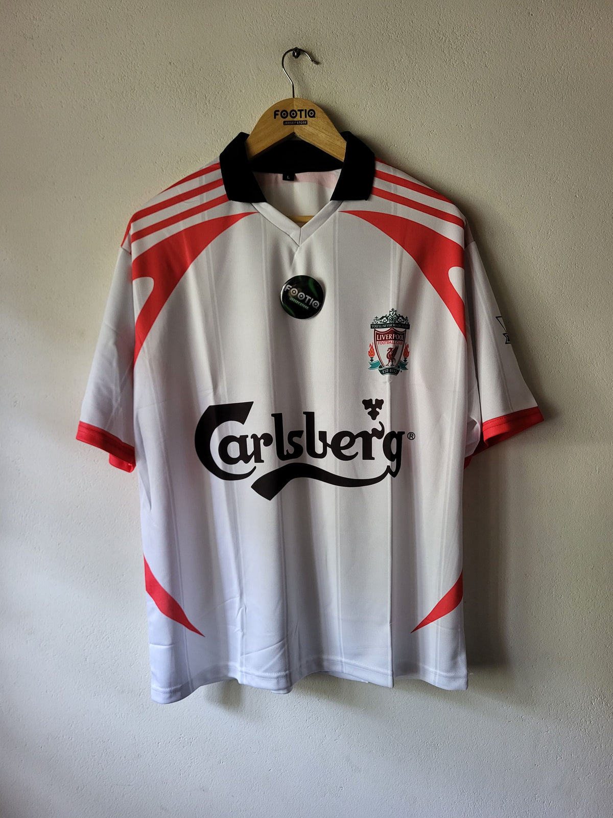 A vintage white Liverpool football jersey with red shoulder stripes and a black collar, hanging on a wooden hanger against a plain wall. The jersey features the Carlsberg sponsor logo on the front.