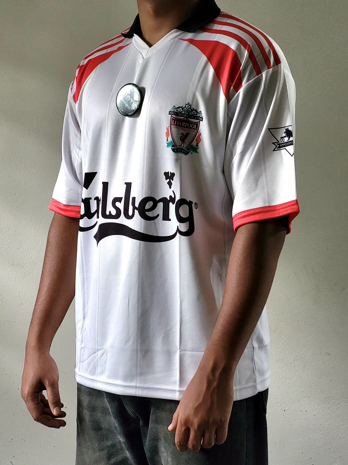 A white short-sleeve soccer jersey with red shoulder stripes, a black collar, and a black Carlsberg sponsor logo, worn by a model. The jersey features the Liverpool club crest on the chest.