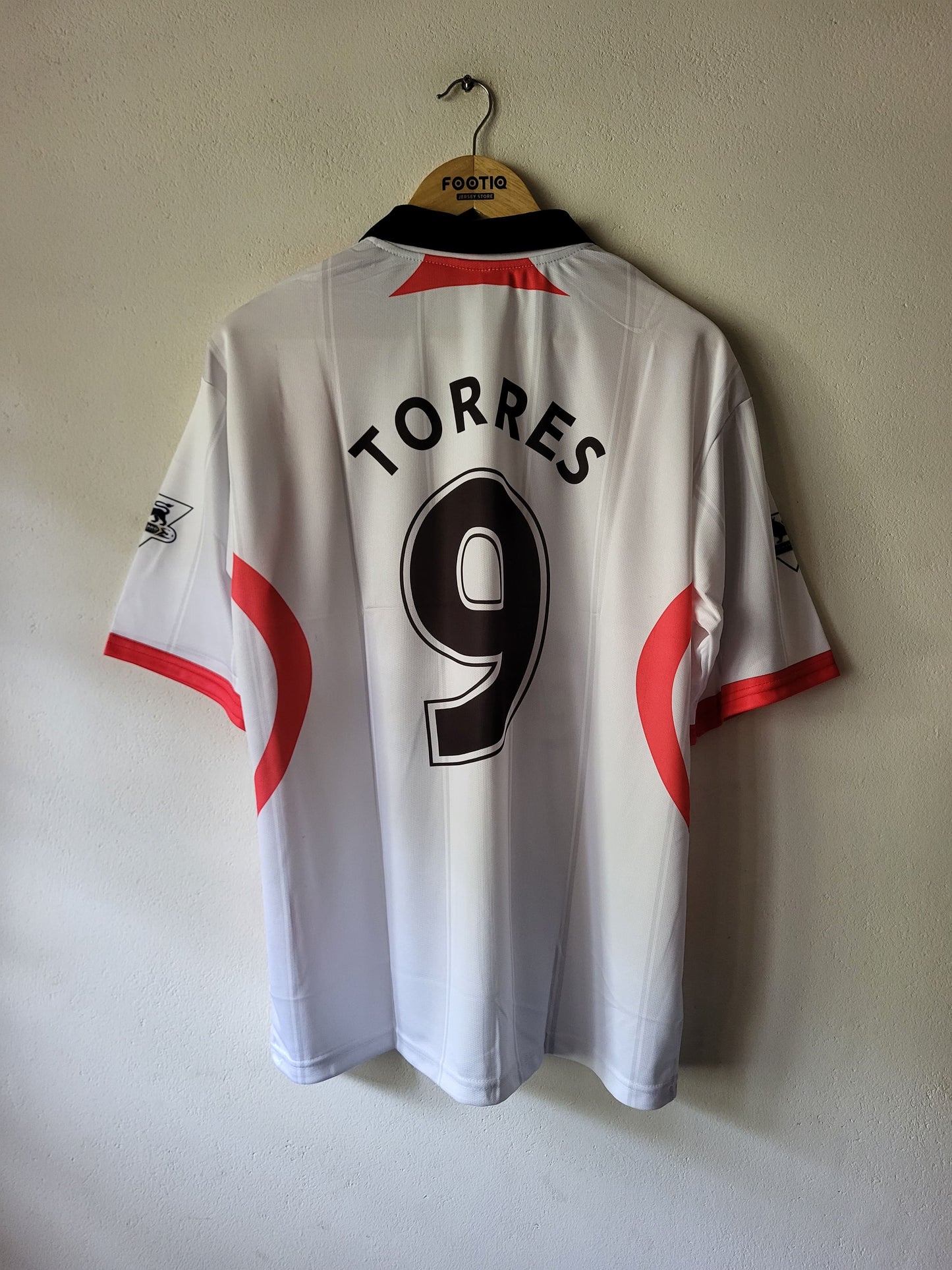 White short-sleeve soccer jersey hanging on a wooden hanger against a plain wall, with the back showing 'TORRES' and the number 9 in black, plus red side accents.