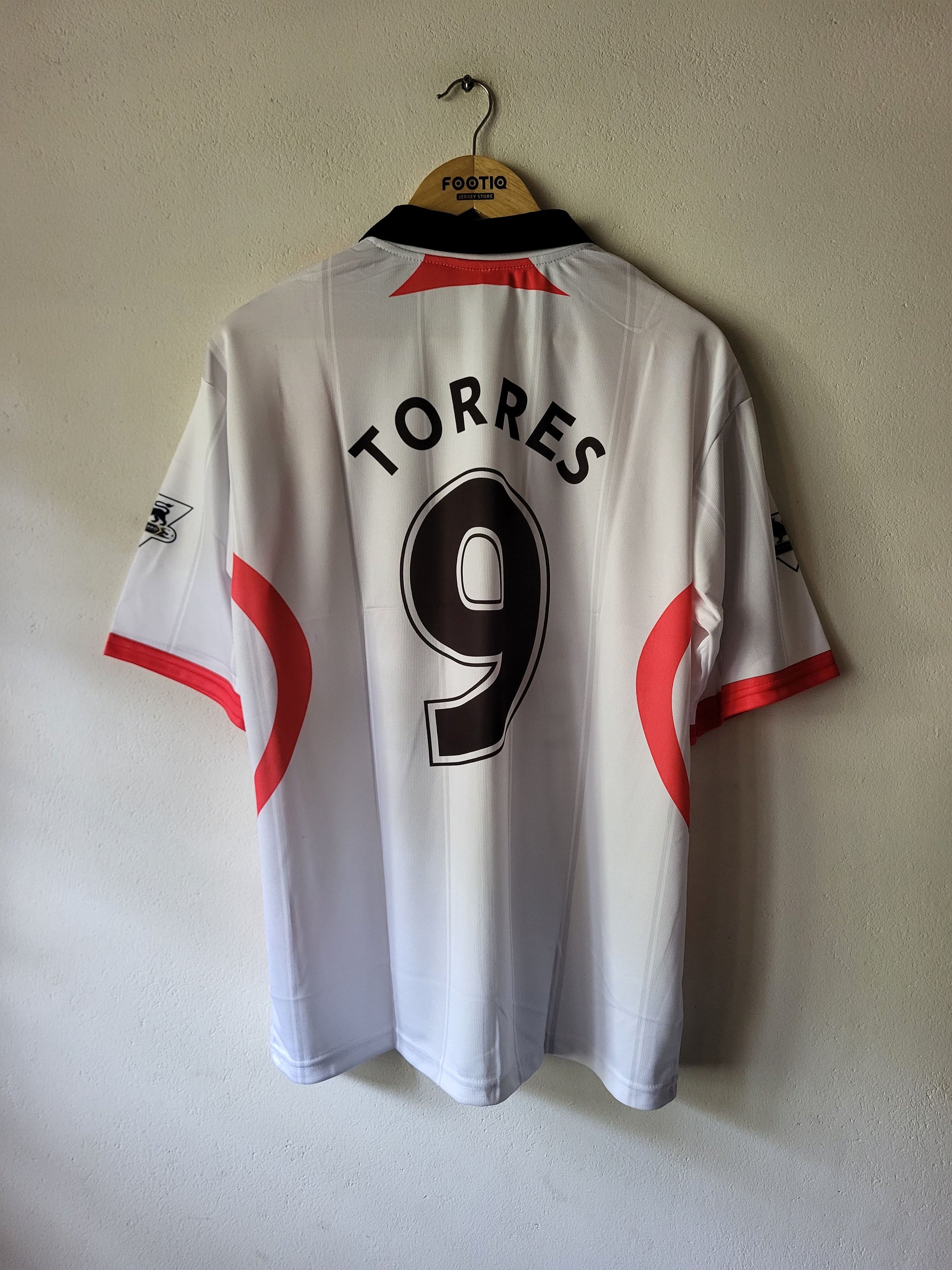 White short-sleeve soccer jersey hanging on a wooden hanger against a plain wall, with the back showing 'TORRES' and the number 9 in black, plus red side accents.