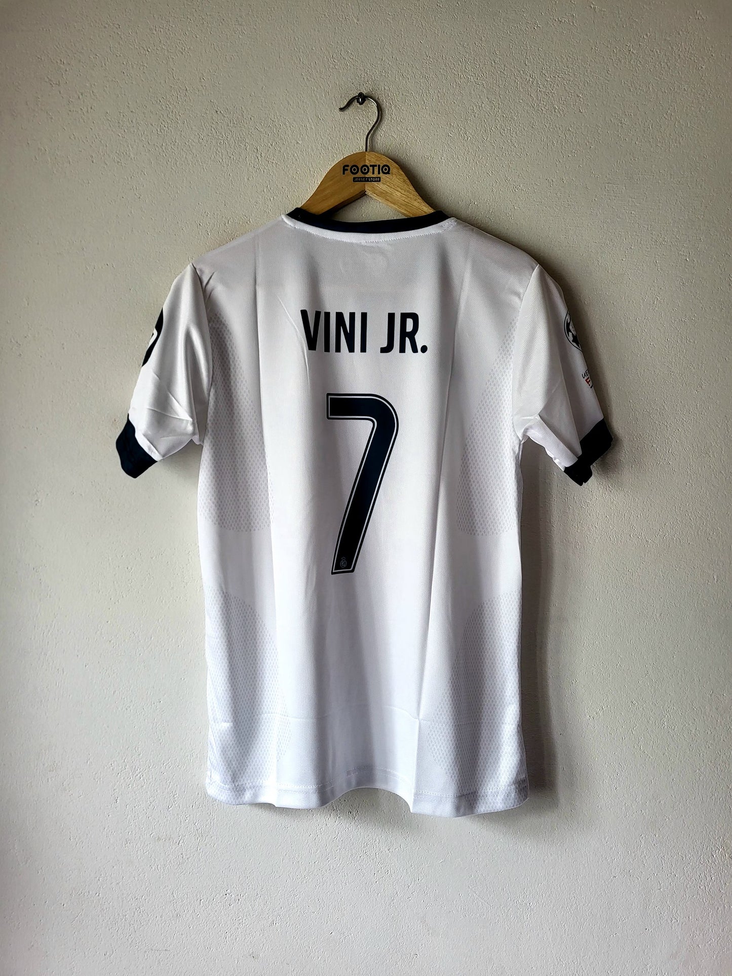 White soccer jersey with 'VINI JR.' and the number 7 printed in black on the back, hanging on a wooden hanger against a textured off-white wall.