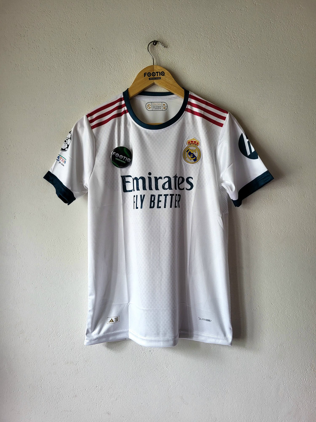 A white Real Madrid soccer jersey with dark navy trim and red shoulder stripes, hanging on a wooden hanger against a light beige wall. The jersey features the Emirates sponsor logo and the club crest.