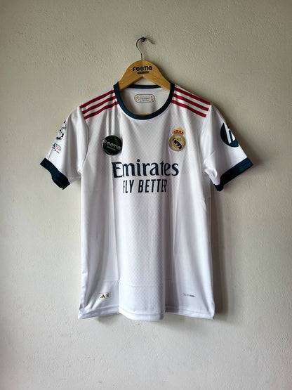 A white Real Madrid soccer jersey with dark navy trim and red shoulder stripes, hanging on a wooden hanger against a light beige wall. The jersey features the Emirates sponsor logo and the club crest.