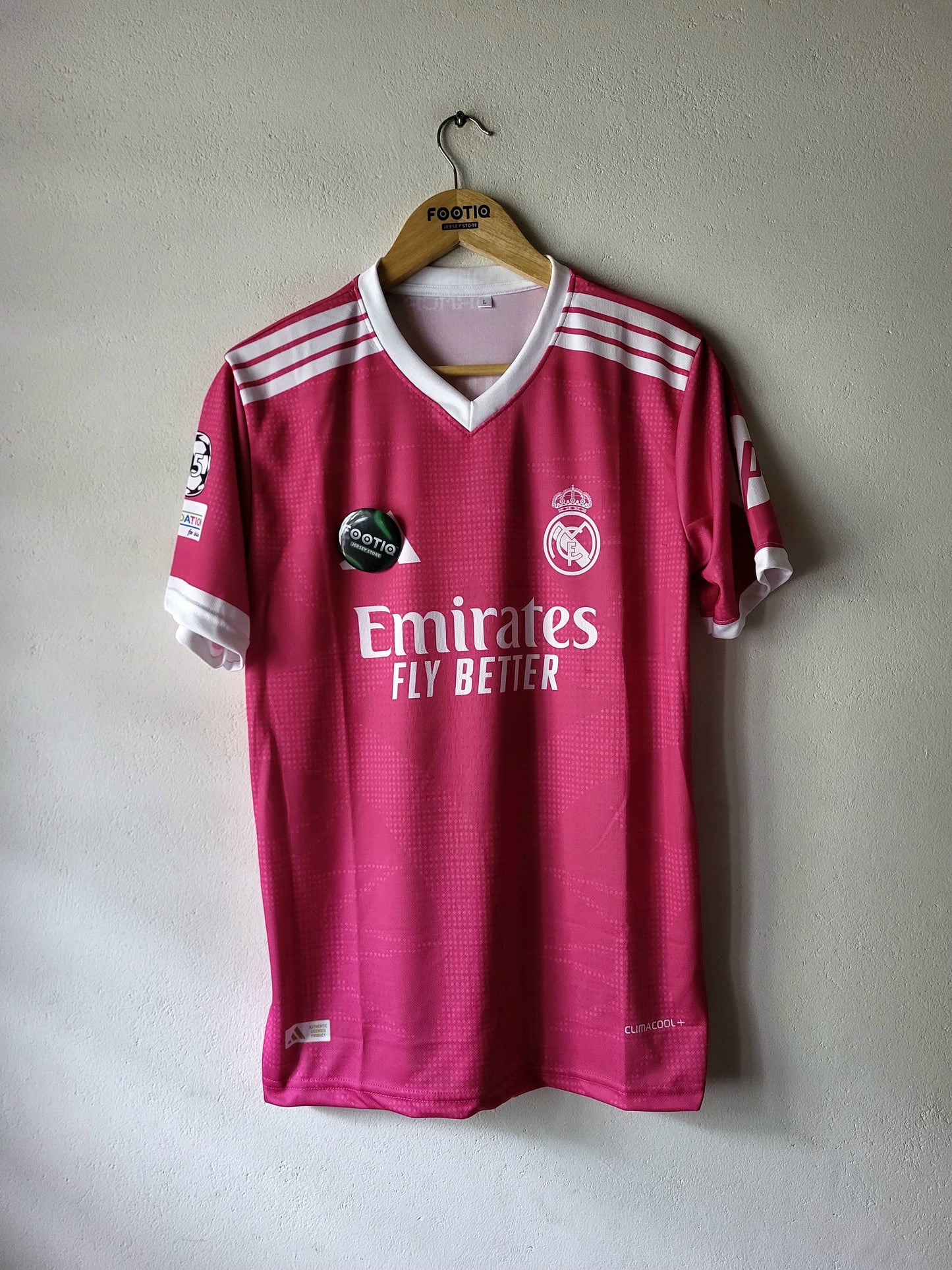 A pink Real Madrid soccer jersey with white accents, hanging on a wooden hanger against a light gray wall. The jersey features the Emirates Fly Better sponsor logo and the Real Madrid club crest.