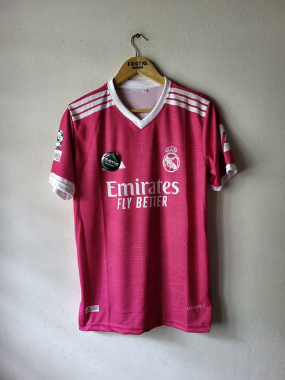 A pink Real Madrid soccer jersey with white accents, hanging on a wooden hanger against a light gray wall. The jersey features the Emirates Fly Better sponsor logo and the Real Madrid club crest.