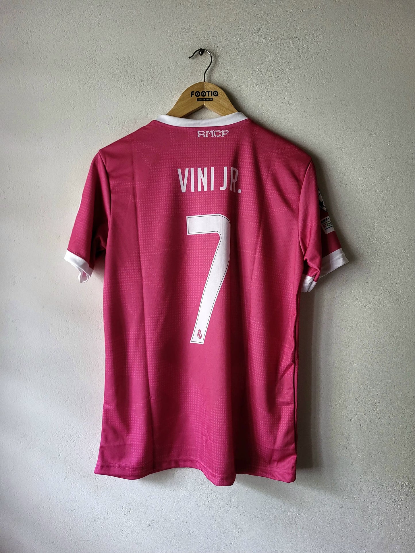 A pink  Real Madrid CF soccer jersey with 'VINI JR.' and the number '7' printed on the back, hanging on a wooden hanger against a textured gray wall.