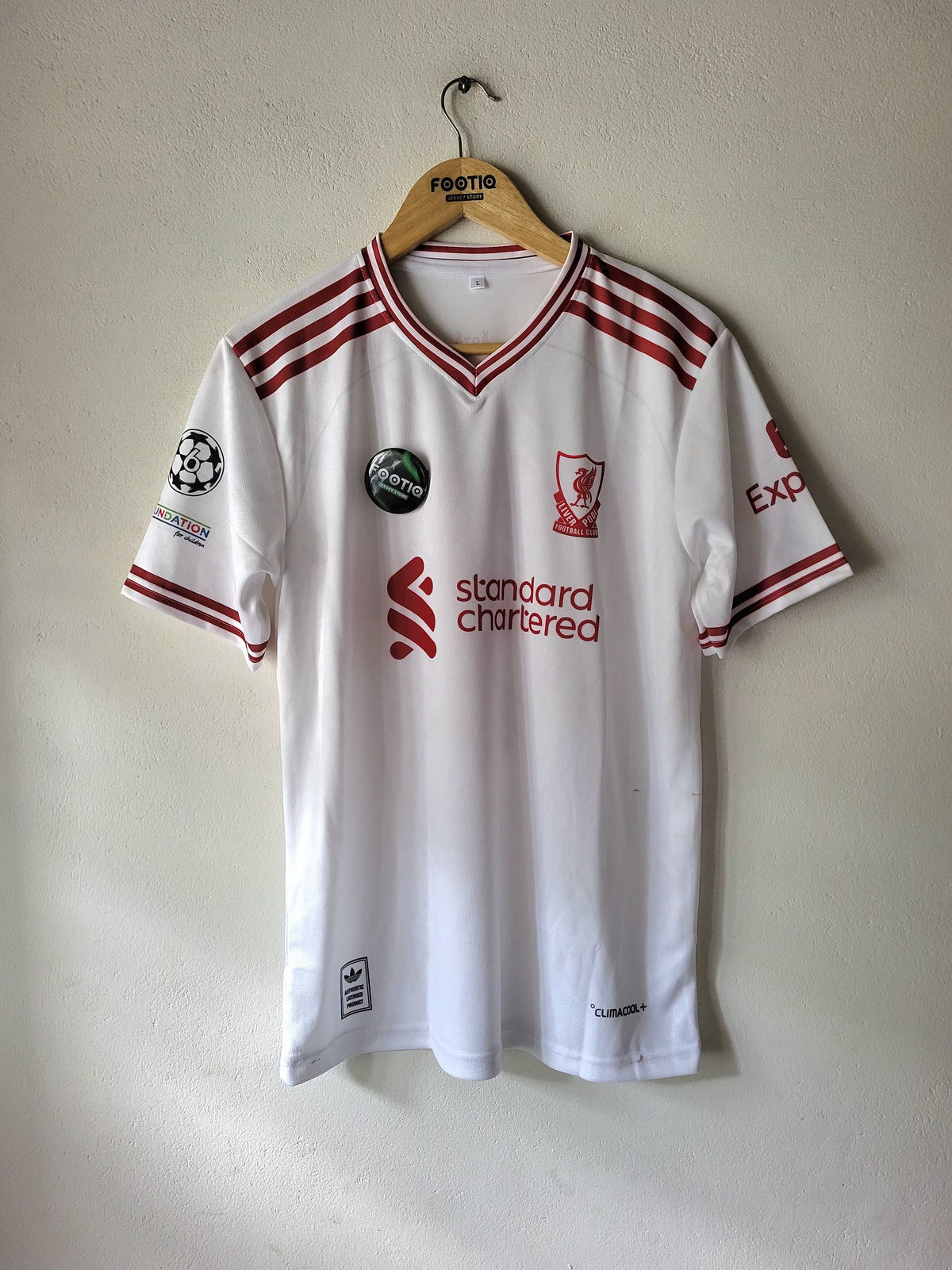 A white Liverpool football jersey with red stripes and logos, hanging on a wooden hanger against a plain wall.