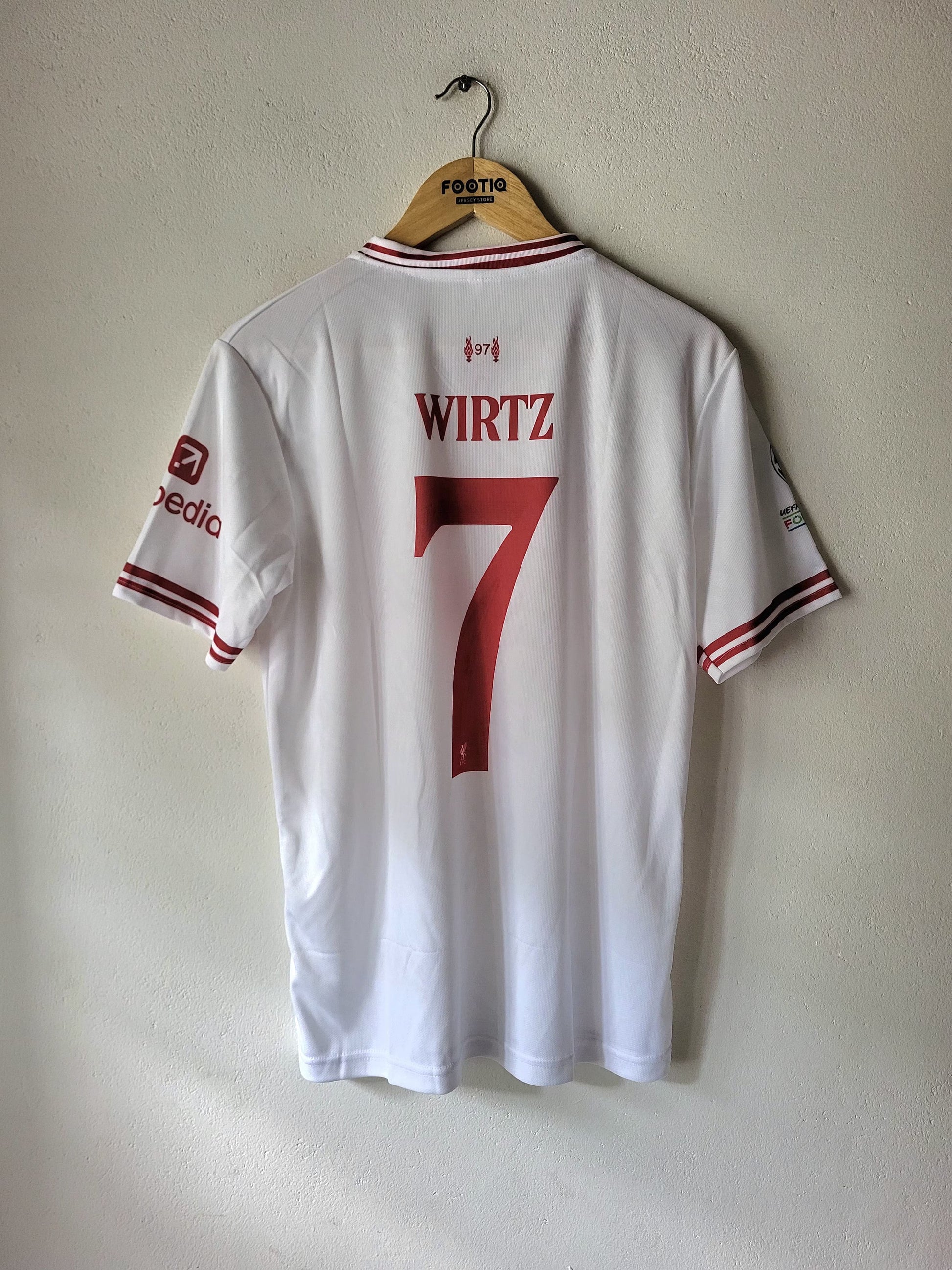 A white Adidas soccer jersey with 'WIRTZ' and the number '7' printed in red on the back, hanging on a wooden hanger against a textured beige wall.