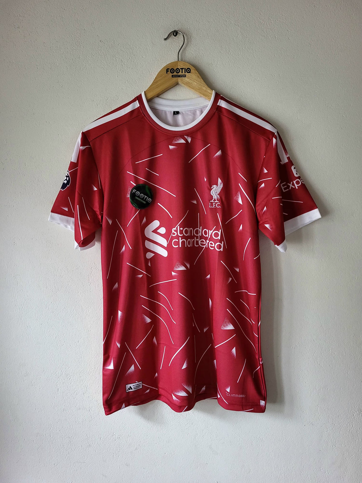 A red and white Liverpool football jersey hanging on a wooden hanger against a plain wall. The shirt features the club's crest, the 'standard Chartered' sponsor logo, and a black circular 'Just Play' badge on the chest.