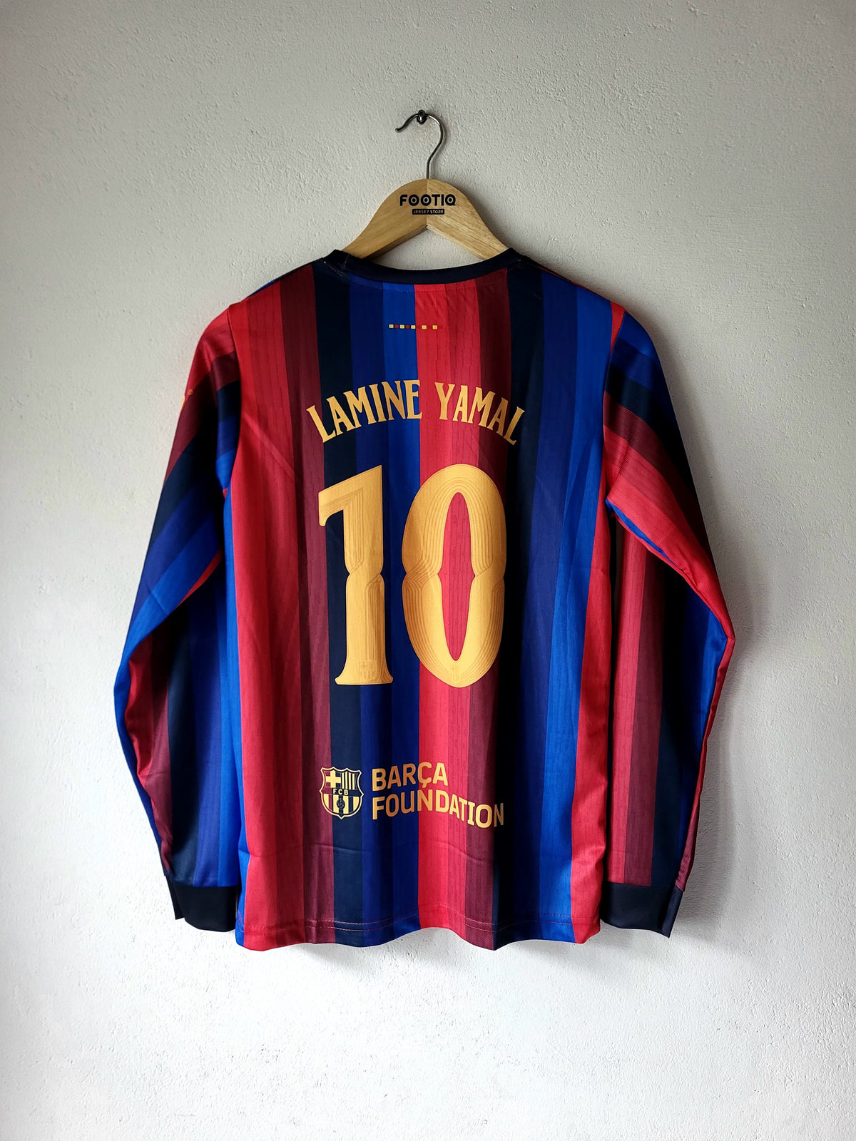 A long-sleeve FC Barcelona football jersey with the back of the number 10 'Lamine Yamal' printed in gold, hanging on a wooden hanger against a white wall.