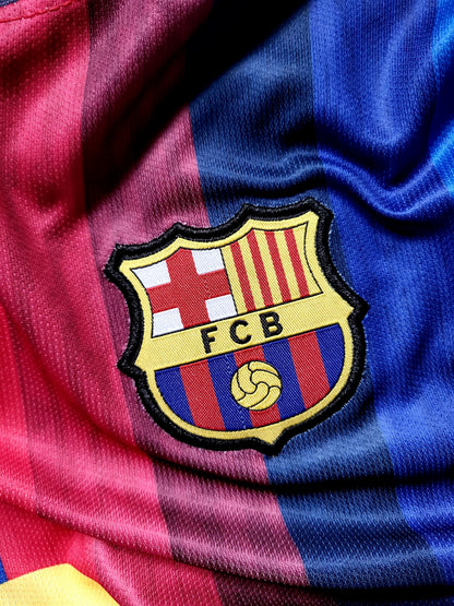 Close-up of an FC Barcelona embroidered crest patch on a satin-textured red and blue striped soccer jersey.