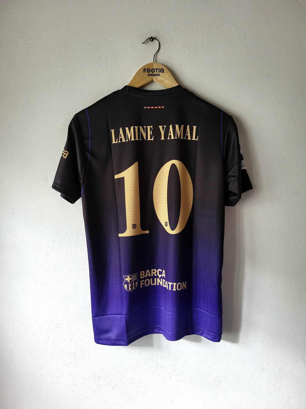 A black and purple gradient soccer jersey hanging on a wooden hanger against a white wall. The back of the jersey features the name 'LAMINE YAMAL' and number '10' in gold, with 'BARCEL ON DEMAND' printed near the bottom.