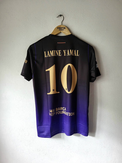 A black and purple gradient soccer jersey hanging on a wooden hanger against a white wall. The back of the jersey features the name 'LAMINE YAMAL' and number '10' in gold, with 'BARCEL ON DEMAND' printed near the bottom.